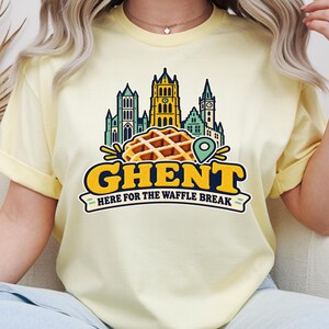 Könnte beinhalten: Ein hellgelbes T-Shirt mit kurzen Ärmeln und einer Grafik der Skyline von Gent, einer goldbraunen Waffel und einer grünen Standortnadel. Der Text lautet "GHENT" in großen gelben Buchstaben und "HERE FOR THE WAFFLE BREAK" darunter.