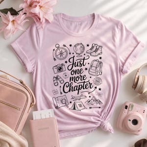 Puede incluir: Una camiseta rosa claro con un estampado gráfico negro con el texto "Just one more Chapter" rodeado de ilustraciones de una brújula, un globo terráqueo, botas de senderismo, mochila, cámara, binoculares, avión, tienda de campaña, montañas y un mapa.