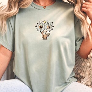 May include: A sage green short-sleeve t-shirt featuring a central graphic of a small robot standing beneath a network of question marks, a lightbulb, and two circles labelled A and B. The robot holds a small sign with the letters A/B printed on it.