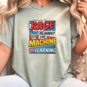 May include: A sage green t-shirt featuring a comic book style graphic with the text "RAGE AGAINST THE MACHINE LEARNING". The design includes a small robot icon and a pink brain illustration set against blue, red, and yellow comic book panels.