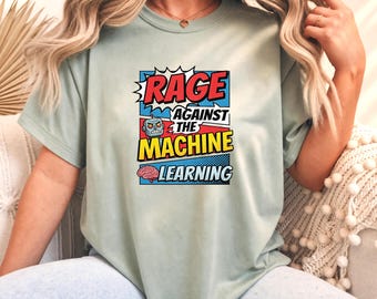 May include: A sage green t-shirt featuring a comic book style graphic with the text "RAGE AGAINST THE MACHINE LEARNING". The design includes a small robot icon and a pink brain illustration set against blue, red, and yellow comic book panels.