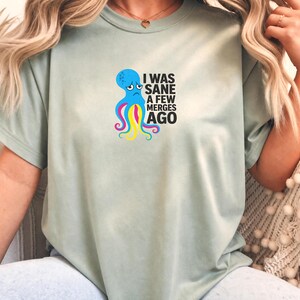 May include: A sage green short-sleeved t-shirt featuring a graphic of a blue octopus with colourful tentacles and the black text "I WAS SANE A FEW MERGES AGO" printed on the front. The shirt has a crew neckline and a relaxed, casual fit.