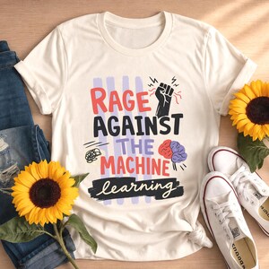 May include: A cream-colored t-shirt featuring a graphic with the text "RAGE AGAINST THE MACHINE learning" in red, black, and white. The design includes a black raised fist icon, a stylized brain, and abstract scribbles over light purple vertical stripes.