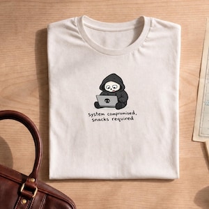 Pode incluir: Uma t-shirt de cor creme com um gráfico de uma personagem de desenho animado com um capuz preto sentada num portátil com um ícone de caveira. Abaixo do gráfico, o texto diz: system compromised, snacks required. Gola redonda.
