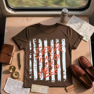 May include: A brown short-sleeved t-shirt featuring a graphic of vertical white distressed stripes and the orange text "EVERY PR IS A CRY FOR HELP." The shirt is displayed alongside a leather notebook, a pen, a compass, a map, and brown loafers.