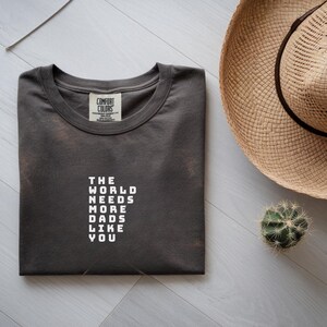 Peut inclure: Un t-shirt à col rond gris anthracite foncé avec une étiquette Comfort Colors à l'intérieur du col. Le devant du t-shirt présente des lettres blanches en bloc qui indiquent THE WORLD NEEDS MORE DADS LIKE YOU, imprimées en format empilé.