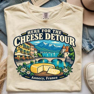 Puede incluir: Una camiseta de color crema con un estampado gráfico que incluye el texto "HERE FOR THE CHEESE DETOUR" y "ANNECY, FRANCE". La ilustración muestra una pintoresca ciudad junto al lago con montañas, un puente de piedra y un queso amarillo.
