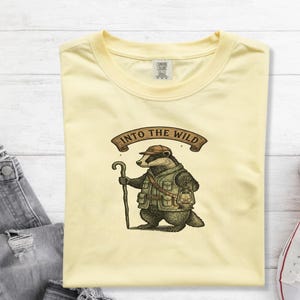 Puede incluir: Una camiseta de color amarillo pálido con una ilustración gráfica de un tejón vestido de explorador, con gorra y chaleco, sosteniendo un bastón de madera y una linterna. Sobre el tejón, una pancarta marrón muestra el texto INTO THE WILD.