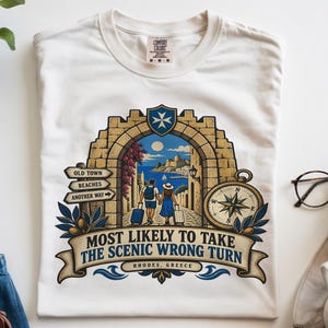 May include: A white t-shirt featuring a graphic of a stone archway framing a coastal scene in Rhodes, Greece. The design includes two travelers with luggage, a compass, and the text "MOST LIKELY TO TAKE THE SCENIC WRONG TURN" on a banner.