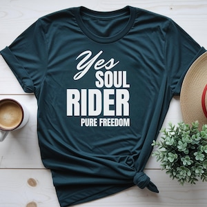 Puede incluir: Una camiseta de manga corta en color verde azulado oscuro con el texto en blanco Yes SOUL RIDER PURE FREEDOM. La prenda tiene un cuello redondo y está anudada en la parte inferior derecha, mostrando un diseño sencillo para uso diario.
