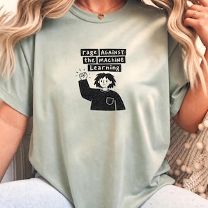 Op de afbeelding: Een licht saliegroen T-shirt met korte mouwen en een zwarte grafische print van een persoon met warrig haar die een vuist omhoog houdt. De tekst boven de figuur luidt "rage AGAINST the MACHINE Learning" in een handgetekend lettertype.