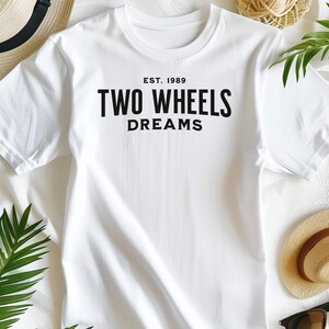Puede incluir: Una camiseta blanca de manga corta con texto negro centrado en el pecho que dice EST. 1989 TWO WHEELS DREAMS. La camiseta está hecha de una tela lisa y se muestra extendida, destacando la tipografía negra y llamativa impresa en la parte frontal.
