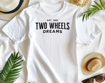 May include: A white short-sleeved t-shirt featuring black text centered on the chest that reads EST. 1989 TWO WHEELS DREAMS. The shirt is made of a smooth fabric and is displayed flat, showcasing the bold, sans-serif typography printed on the front.