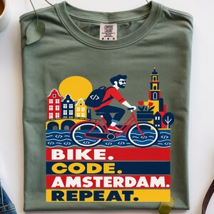 Puede incluir: Una camiseta verde oliva con un gráfico de una persona con barba y mochila montando una bicicleta roja por un paisaje urbano de Ámsterdam. El diseño incluye un sol amarillo, un canal y edificios. El texto dice: BIKE. CODE. AMSTERDAM. REPEAT.