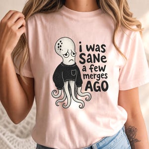 Op de afbeelding: Een lichtroze T-shirt met korte mouwen en een zwart-witte illustratie van een verdrietige octopus in een zwarte trui. De tekst die in het zwart naast de octopus is gedrukt, luidt "i was SaNe a few merges AGO" in een mix van kleine en hoofdletters.