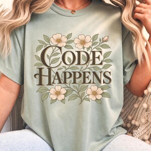 May include: A light sage green t-shirt featuring the text "CODE HAPPENS" in a dark brown serif font. The text is surrounded by a botanical illustration of light pink flowers with yellow centres and green leaves arranged in a rectangular frame.