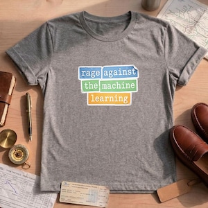 May include: A heather grey short-sleeved t-shirt featuring a graphic with three stacked rectangular blocks in blue, green, and yellow. The text reads "rage against the machine learning" in a white, typewriter-style font across the coloured blocks.