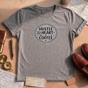 Può includere: Una t-shirt grigio melange a maniche corte con una grafica circolare nera sul petto. Il design presenta il testo "HUSTLE & HEART & COFFEE" in un carattere grassetto senza grazie, circondato da un bordo decorativo di chicchi di caffè e stelle.