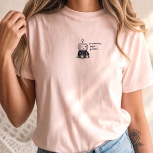 Op de afbeelding: Een lichtroze T-shirt met korte mouwen en een kleine afbeelding op de linkerborst van een zittende robot met een gloeilamp boven zijn hoofd. De tekst naast de robot luidt "processing... logic... ponder..." in een eenvoudig zwart lettertype.