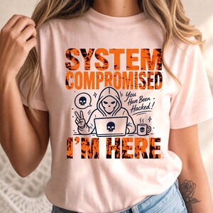 Peut inclure: Un t-shirt rose clair avec une impression graphique portant le texte "SYSTEM COMPROMISED" et "I'M HERE" en lettres orange usées. Le motif inclut une silhouette à capuche devant un ordinateur, une icône de crâne et le texte "You Have Been Hacked!"