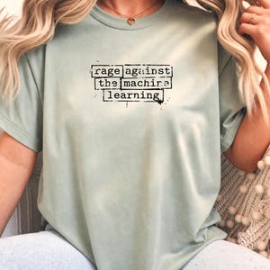 May include: A sage green short-sleeve t-shirt featuring black text printed in a typewriter-style font within rectangular boxes. The text reads "rage against the machine learning" with a distressed, ink-stamped appearance on the front chest area.