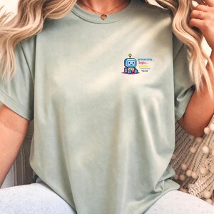 Op de afbeelding: Een saliegroen t-shirt met korte mouwen en een kleine afbeelding op de linkerborst. De afbeelding toont een blauwe en roze robot met een geel gloeilampje boven het hoofd, vergezeld van de tekst "processing... logic... gender".