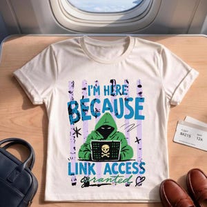 Op de afbeelding: Een crèmekleurig t-shirt met een afbeelding van een figuur met een capuchon die een laptop gebruikt met een schedel- en kruisbeenderenpictogram. De tekst luidt I'M HERE BECAUSE LINK ACCESS GRANTED, met verticale paarse strepen.