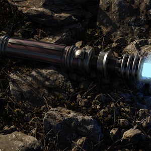 May include: A silver and brown lightsaber with a blue blade, lying on a bed of rocks and dirt.