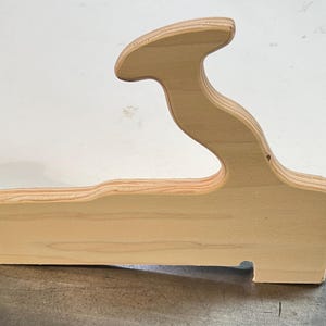 May include: A light-colored wooden handle with a smooth, curved silhouette and visible layered plywood grain. The object features a distinct ergonomic grip shape with a flat base and a rounded top section, showcasing natural wood tones and textures.