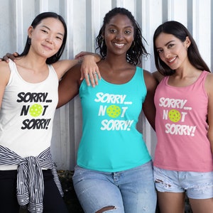 May include: Three women wearing tank tops with the text "SORRY... NOT SORRY!" and a yellow pickleball graphic. The tank tops are white, turquoise, and pink.