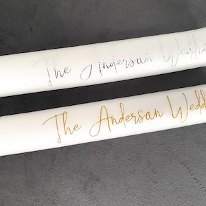 May include: Two white wedding candles with text. One candle has silver text reading "The Simpson Wedding." The other has gold text reading "The Anderson Wedding."