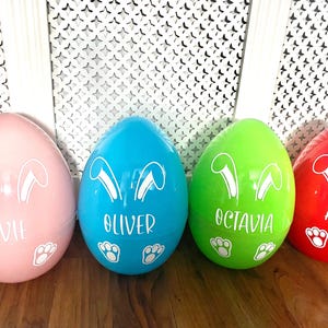 Puede incluir: Cuatro huevos de Pascua grandes y coloridos en rosa, azul, verde y rojo. Cada huevo presenta orejas de conejo blancas, huellas de patas y un nombre: Evie, Oliver, Octavia y Ava. Una decoración festiva de Pascua.