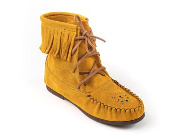 May include: Mustard yellow suede moccasin boot with fringe detail. The boot has brown laces and decorative beading on the toe. The sole is a dark brown colour. A stylish and comfortable footwear option.