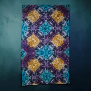 May include: A rectangular fabric piece featuring a tie-dye pattern with repeating geometric shapes in shades of deep purple, bright turquoise, and golden yellow. The design displays a symmetrical, kaleidoscopic arrangement of colors.