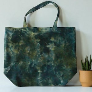 May include: A large tote bag with a tie-dye pattern in shades of green and blue. The bag has two handles and is made of a soft, flexible material. A small potted plant is next to the bag.