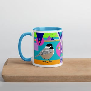 May include: A white ceramic mug with a blue interior and handle. The mug features a colorful geometric design with a bird illustration. The bird is a chickadee. The mug sits on a wooden surface.