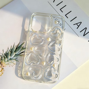 May include: Transparent phone case featuring a heart pattern. The case has raised heart shapes and small, pearl-like embellishments. The clear design allows the phone's colour to be visible.