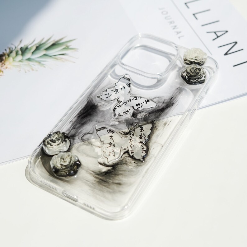 May include: Transparent phone case with butterfly and rose decorations. The case has two butterfly adornments with script and three small rose accents. The background has a black and white marbled effect.
