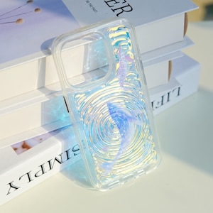 May include: Clear phone case with a whale design. The case has a swirling water effect with a blue and purple whale. The case is made of clear plastic and is designed to protect the phone.