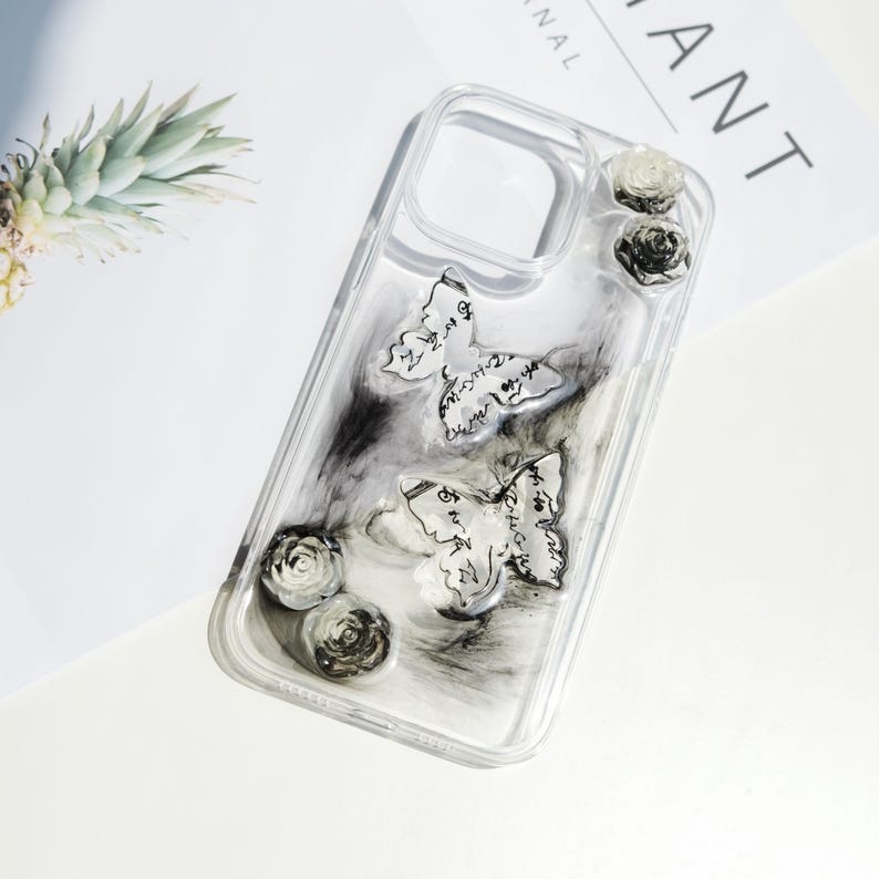 May include: Transparent phone case featuring a black and white marble effect, decorated with two butterfly motifs and three rose details. The butterflies have script writing. The case is designed for a smartphone.