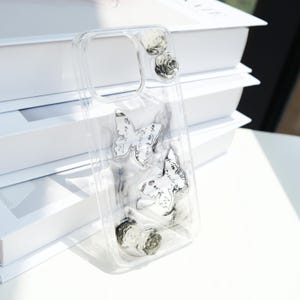 May include: Transparent phone case decorated with silver butterflies and roses. The case has two silver butterflies with script details and small silver roses at the top and bottom. The clear case allows the phone's colour to be visible.