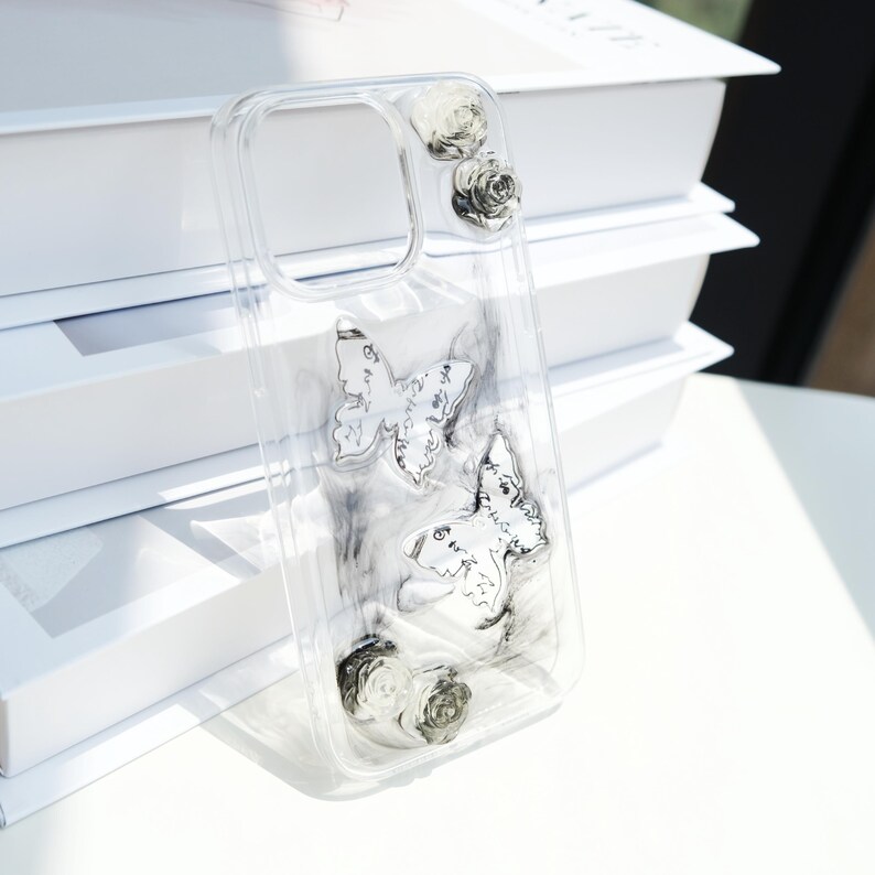 May include: Transparent phone case decorated with silver butterflies and roses. The case has two silver butterflies with script details and small silver roses at the top and bottom. The clear case allows the phone's colour to be visible.