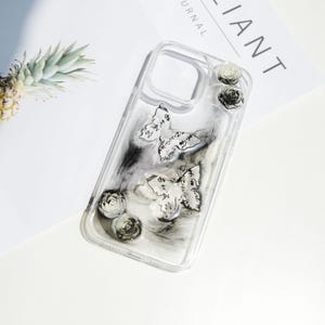 May include: Clear phone case with a marble design, featuring two butterfly accents with script and small rose embellishments. The case has a clear border and a black and white marbled background.