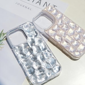 以下が含まれることがあります： 装飾的なデザインの透明な携帯電話ケースが2つ。1つのケースは、銀色のテクスチャ加工された幾何学模様が特徴で、もう1つは真珠のようなボーダーと、同様の、より明るい幾何学模様があります。ケースは白い表面にあります。
