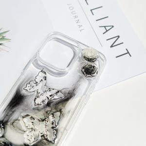 May include: Transparent phone case featuring butterfly motifs and two rose embellishments. The butterflies have script details, set against a black and grey marbled effect. The case is on a white surface.
