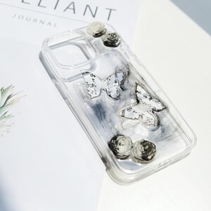 May include: Clear phone case with butterfly and rose embellishments. The case features two butterfly designs with script and several small rose accents. The background has a smoky, marbled effect.