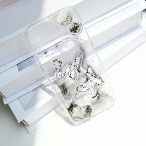 May include: Transparent phone case with butterfly and rose details. The case has two butterfly designs with script writing and small rose embellishments. The case has a clear, transparent design.