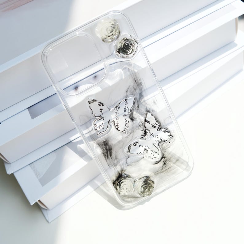 May include: Transparent phone case with butterfly and rose details. The case has two butterfly designs with script writing and small rose embellishments. The case has a clear, transparent design.