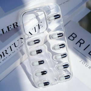 May include: Transparent phone case featuring a pill design. The case is clear with multiple white and black capsule-shaped decorations. The background shows the text "GIVE TIME A LITT".