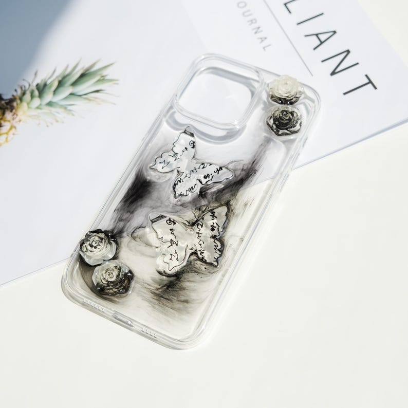 May include: Transparent phone case decorated with butterfly and rose details. The case has two butterflies with script-like patterns and three small roses. The design includes black and white marbled effects, creating a stylish and unique accessory.